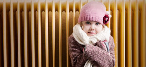 little girl wearing hat and scarf huddled next to a heater